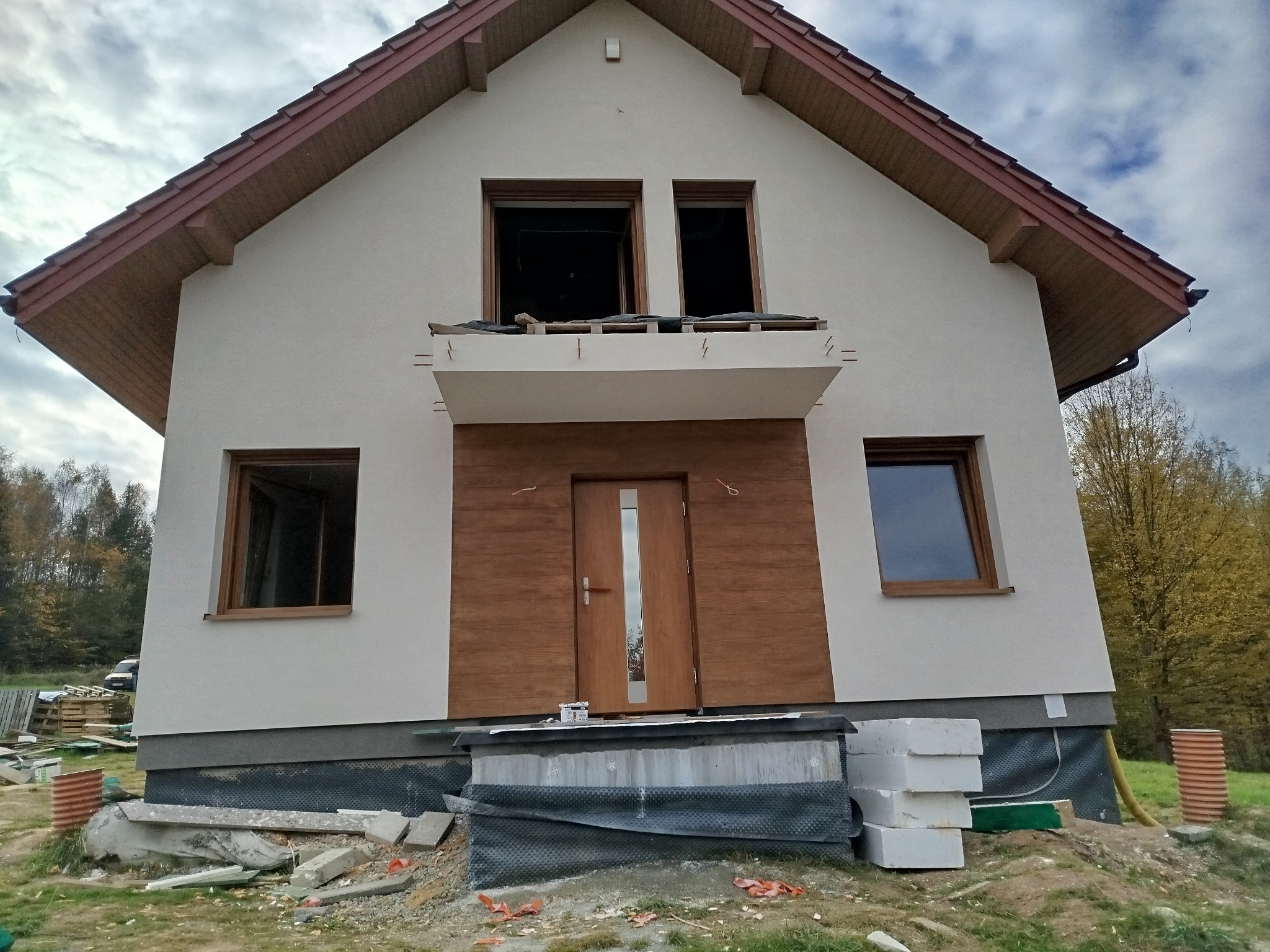 Surowa elewacja domu jednorodzinnego w trakcie wykończenia, z widocznymi oknami bez szyb, drewnianą stolarką drzwiową i fragmentami materiałów budowlanych wokół fundamentów.