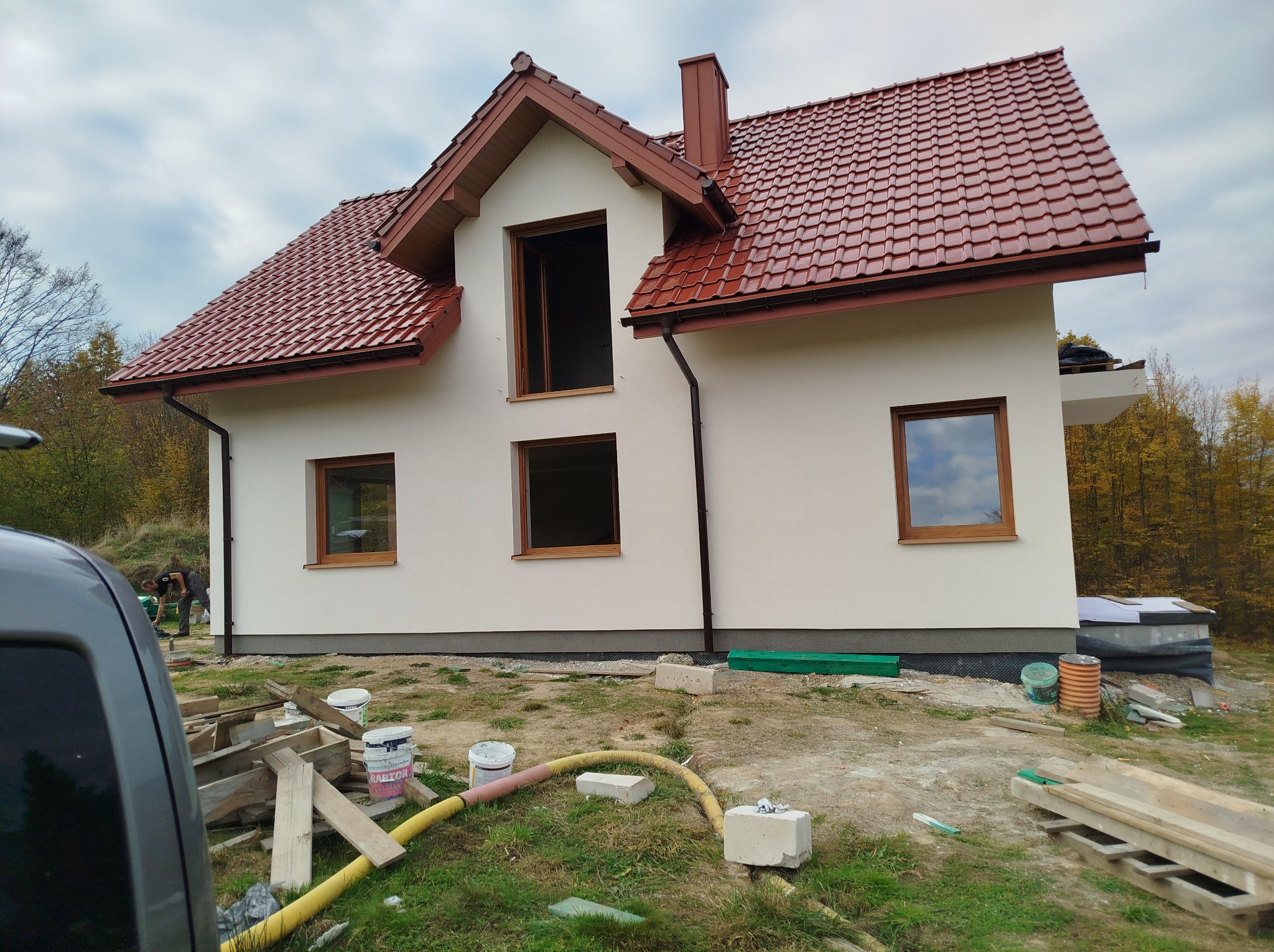 Dwukondygnacyjny dom z czerwonym dachem w trakcie prac wykończeniowych elewacji, widoczne rusztowania, materiały budowlane i narzędzia.