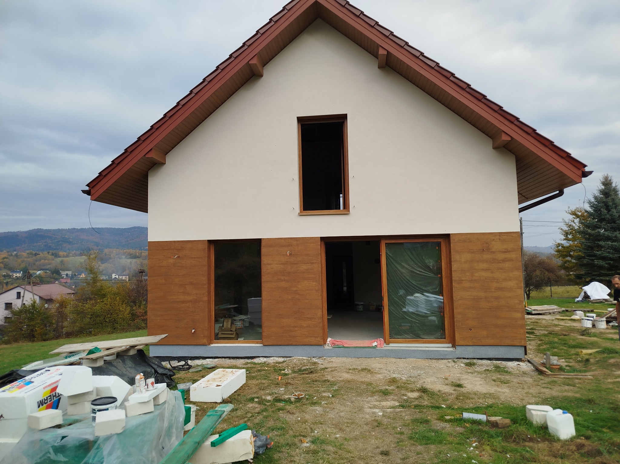 Dwukondygnacyjny dom jednorodzinny z drewnopodobną elewacją w trakcie montażu, widoczne płyty styropianowe i puszki farb na trawniku.