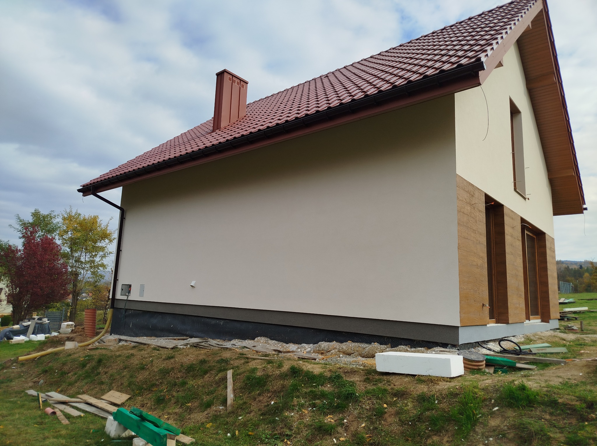 Boczny widok domu z nową, jasną elewacją, brązową dachówką i fragmentem elewacji imitującej drewno. Wokół domu widoczne materiały budowlane i narzędzia.