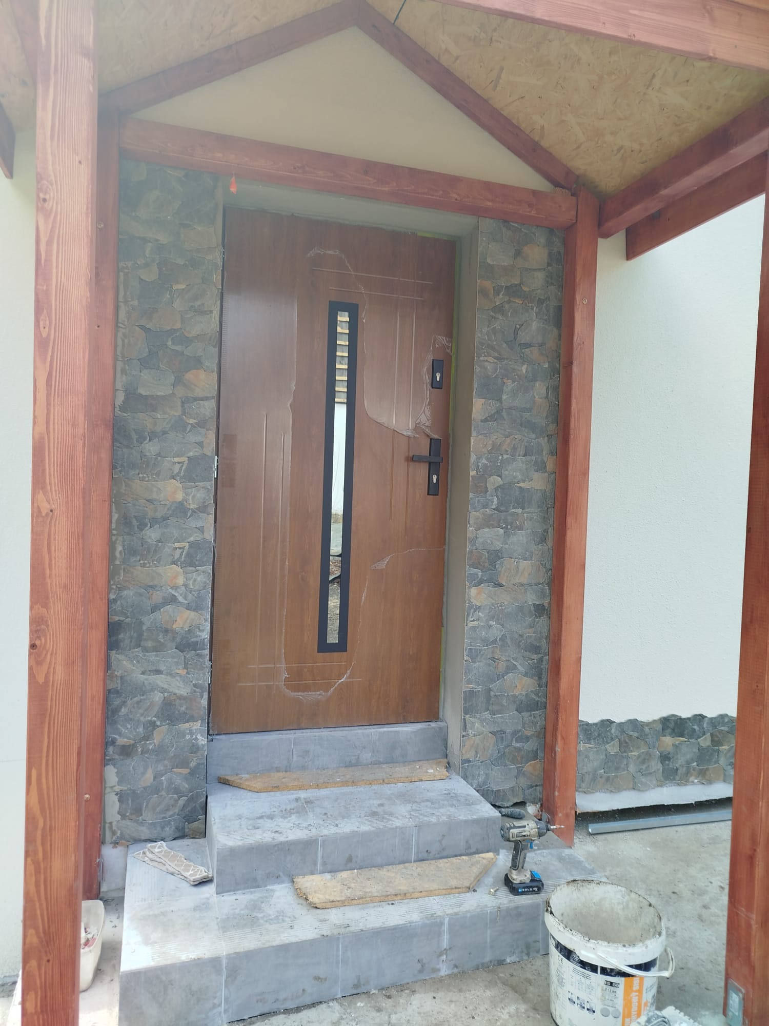 Wykończenie wejścia do domu: obłożona kamieniem elewacja, drewniana konstrukcja zadaszenia, brązowe drzwi wejściowe. Widoczne narzędzia i wiadro farby.