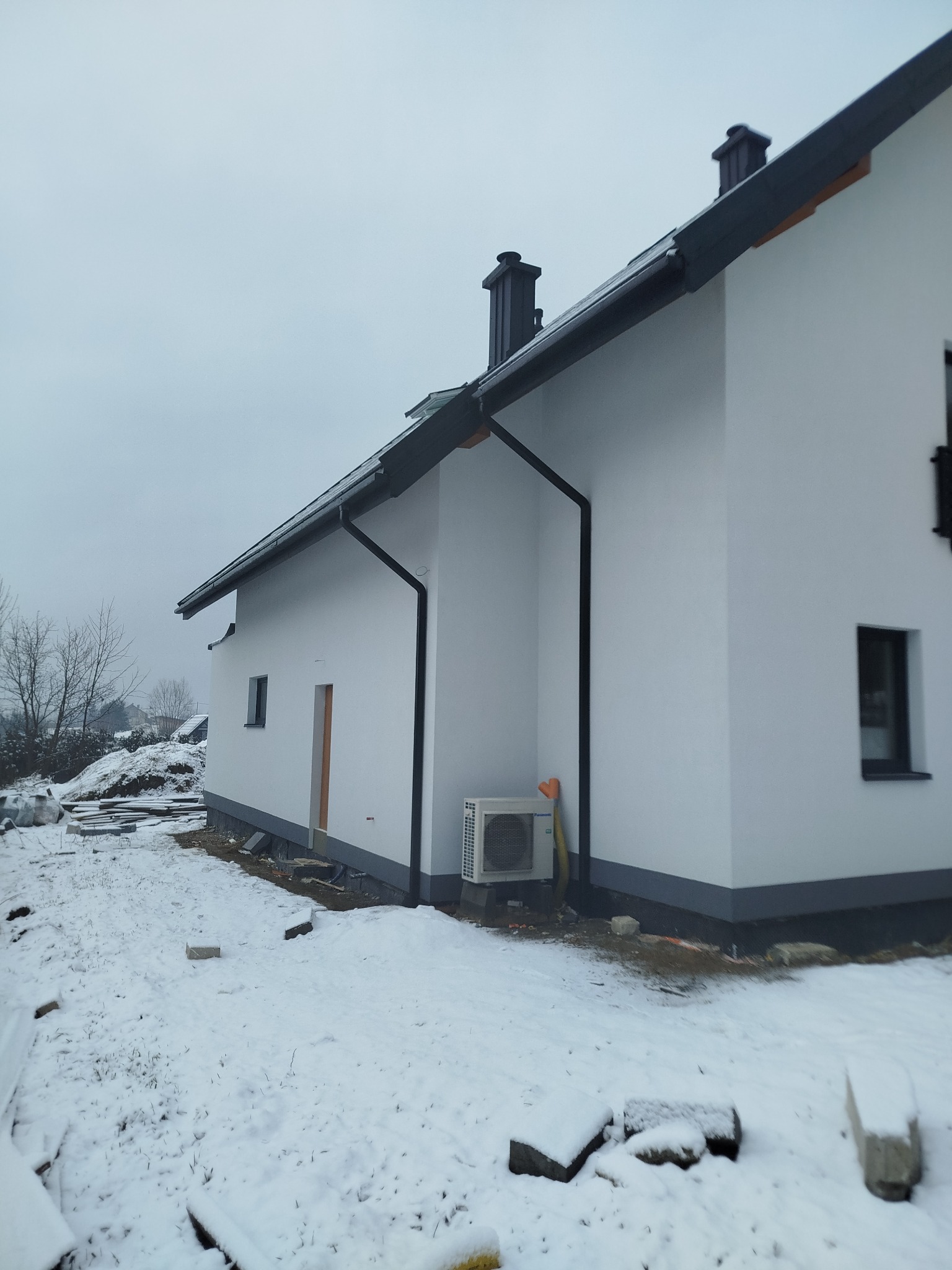 Biały dom z szarymi elementami wykończeniowymi i klimatyzatorem na zewnątrz, otoczony śniegiem w zimowy dzień. Widok z boku, z elementami placu budowy w tle.