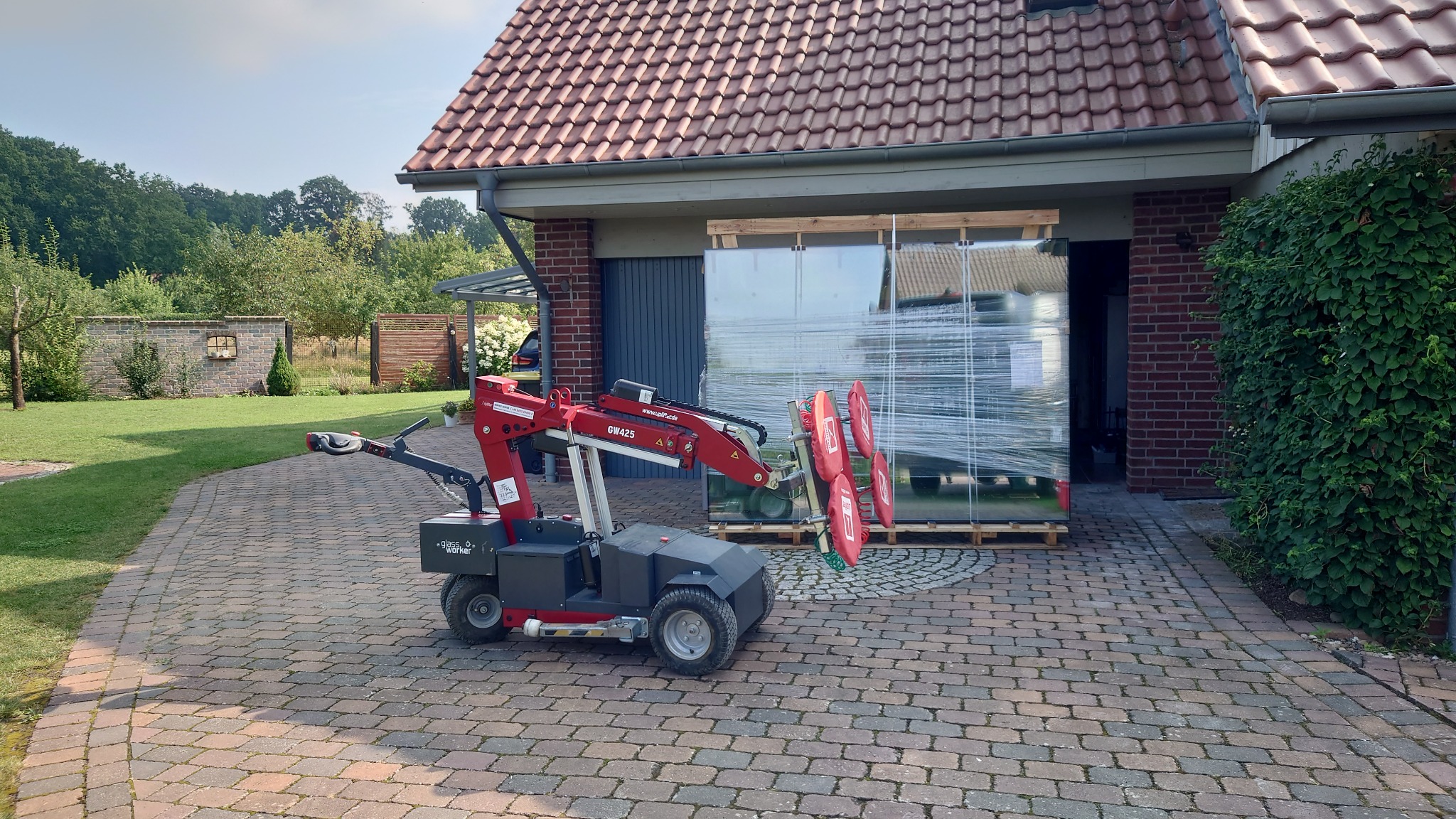 Czerwony podnośnik próżniowy transportuje duże okno owinięte folią stretch na palecie przed garażem w domu jednorodzinnym z czerwoną dachówką.