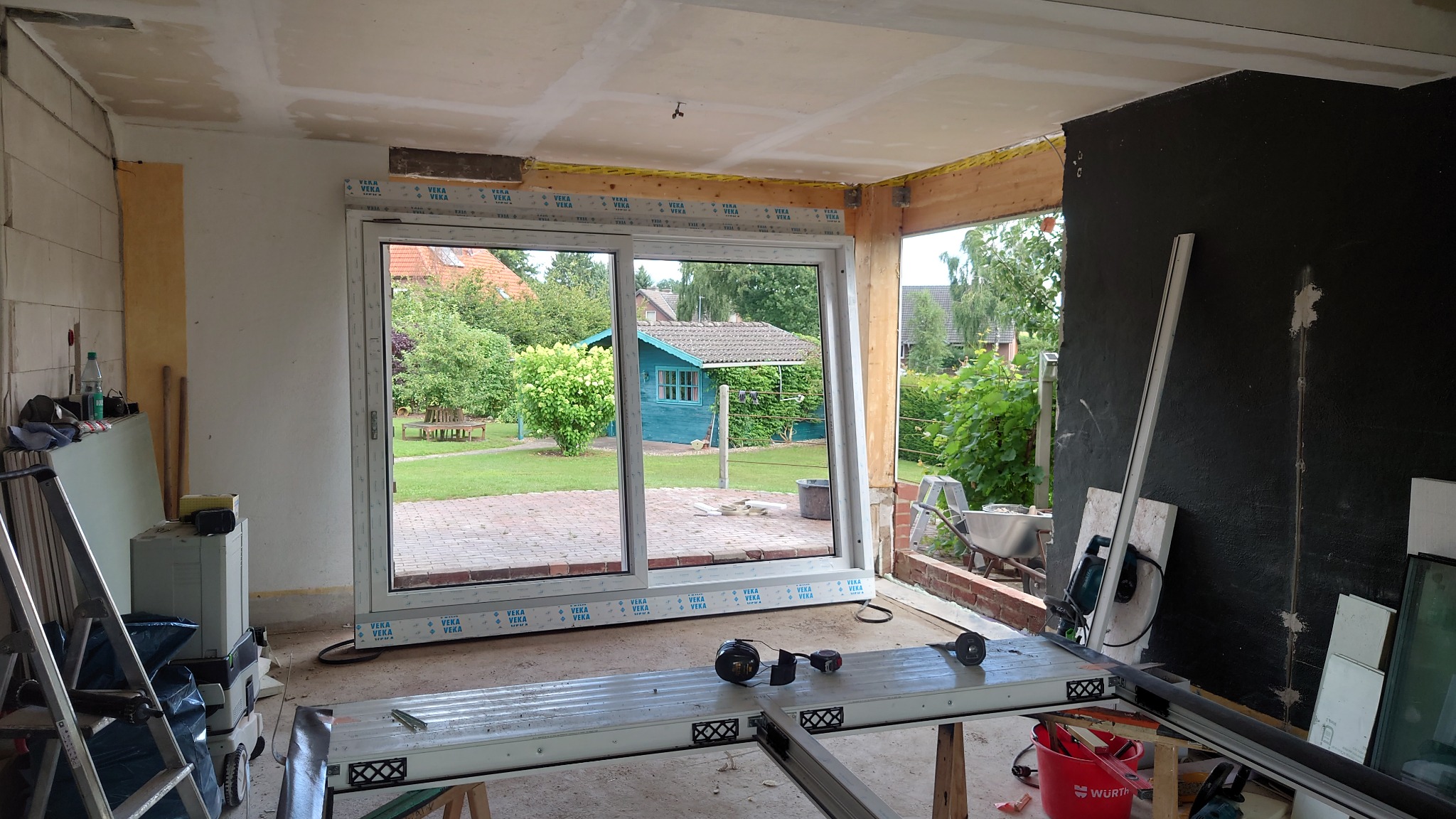 Montaż przesuwnych drzwi balkonowych z białym profilem w surowym stanie wykończenia wnętrza. Widoczne narzędzia, elementy konstrukcyjne i ogród za oknem.