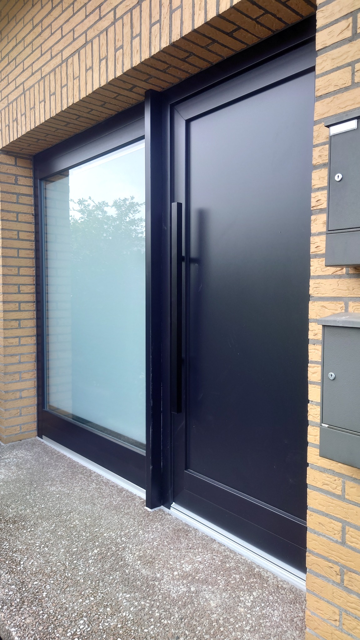 Nowoczesne, czarne drzwi wejściowe z długim, pionowym uchwytem, po lewej stronie przeszklenie z matową szybą, obok ściana z żółtej cegły i skrzynki na listy.
