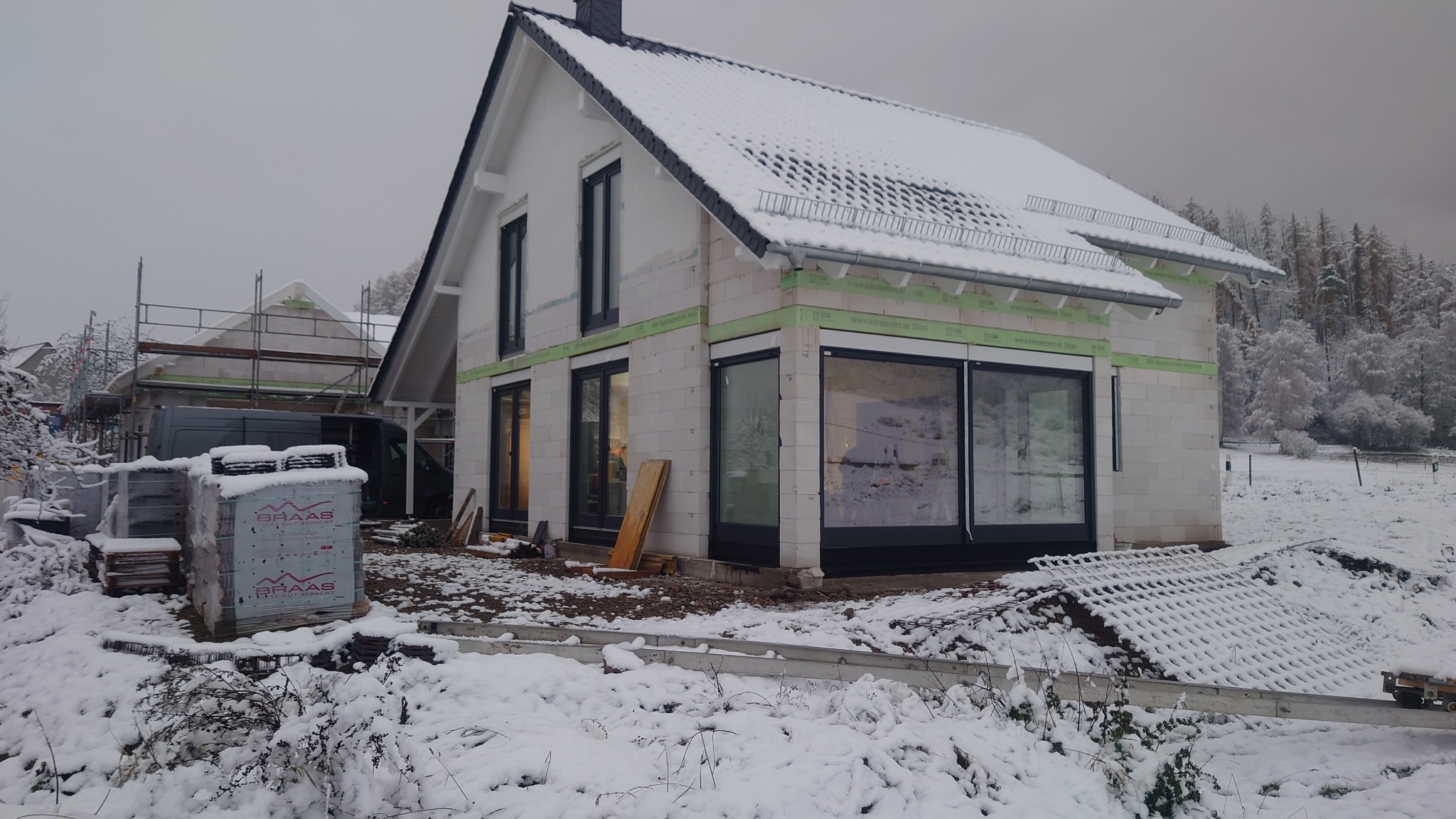 Dom w trakcie budowy zimą, z zamontowanymi oknami PCV i aluminiowymi, widoczne rusztowania i materiały budowlane pokryte śniegiem.