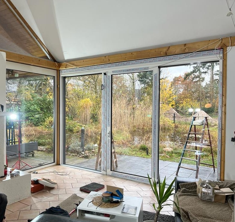 Wnętrze z dużymi przesuwnymi oknami z białymi ramami, widok na ogród z oczkiem wodnym, drabina oparta o ścianę, częściowo wykończone drewniane obramowanie okien.