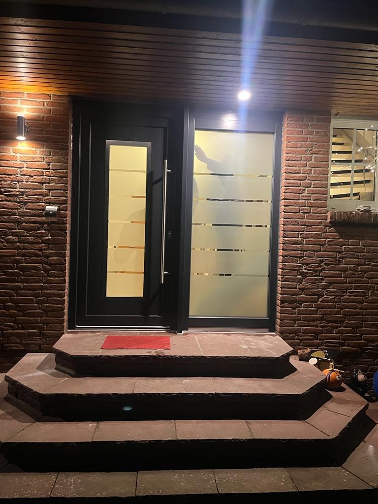 Nowoczesne drzwi wejściowe z przeszkleniem w czarnej ramie, prowadzące do domu z cegły, oświetlone lampami zewnętrznymi i z kilkoma stopniami wejściowymi.