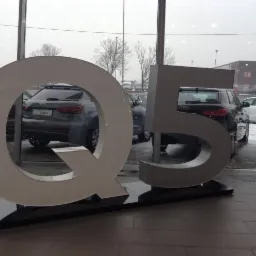 Duży, przestrzenny napis 'Q5' w salonie samochodowym Audi, z widocznymi samochodami marki Audi za szybą