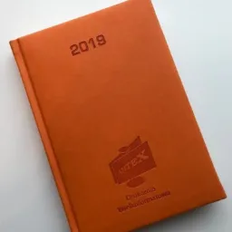 Pomarańczowy kalendarz książkowy z wytłoczonym rokiem 2019 i logo firmy Drukarnia Wielkoformatowa AMTEX leżący na białym tle.
