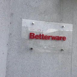 Tablica z logo 'Betterware' z czerwonymi literami na przezroczystym podłożu, zamocowana na szarej, chropowatej ścianie zewnętrznej budynku za pomocą czterech metalowych śrub dystansowych.