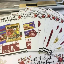 Stos wydrukowanych kartek z świątecznymi motywami i ofertami promocyjnymi na słodycze, ułożonych na stole w drukarni.