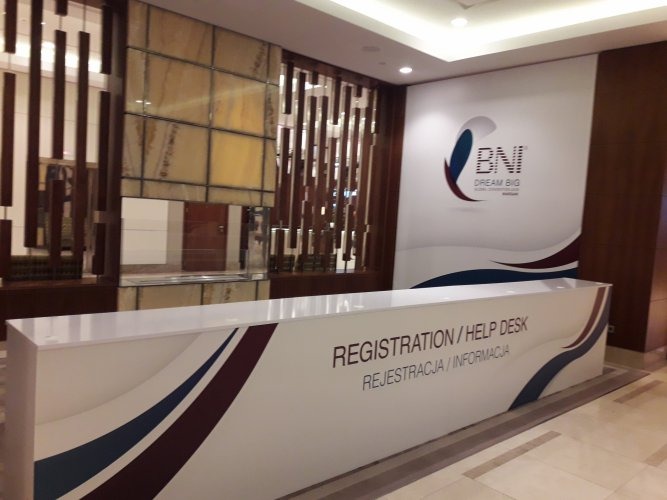 Recepcja z napisem 'REGISTRATION / HELP DESK' i logo BNI na ścianie, widoczne elementy dekoracyjne z drewna i kamienia