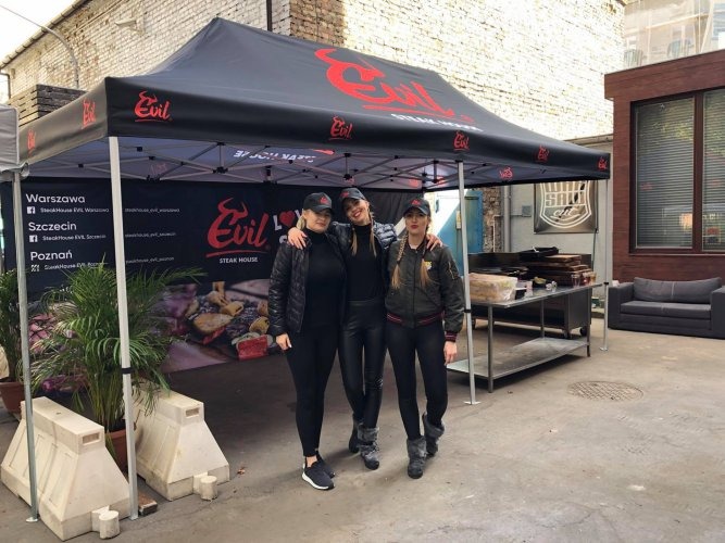 Czarny namiot z czerwonym logo Evil Steak House, trzy kobiety w czapkach z logo firmy stojące przed namiotem, adresy lokali w Warszawie, Szczecinie i Poznaniu na ścianie namiotu.