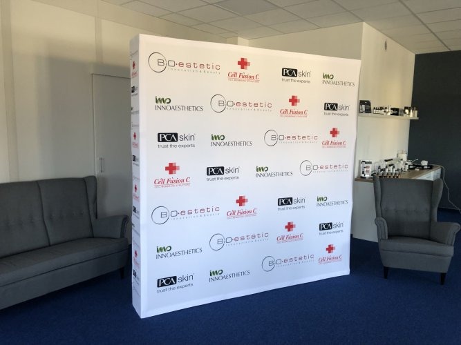 Ścianka reklamowa z powtarzającymi się logotypami firm z branży kosmetycznej, ustawiona w jasnym pomieszczeniu z szarą sofą i fotelem.