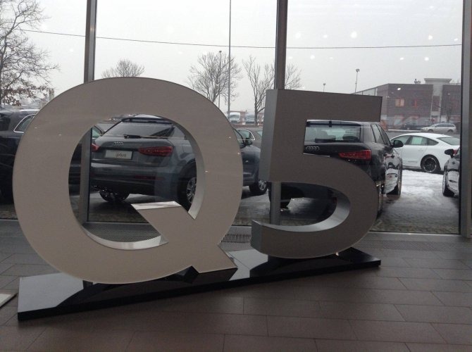 Duży, przestrzenny napis 'Q5' w salonie samochodowym Audi, z widocznymi samochodami marki Audi za szybą
