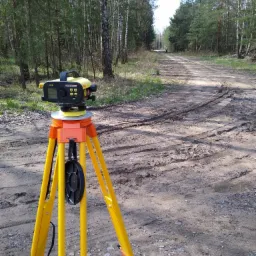 Żółty niwelator Leica Sprinter 150M na statywie, ustawiony na piaszczystej drodze w lesie, w tle widoczna sylwetka osoby.
