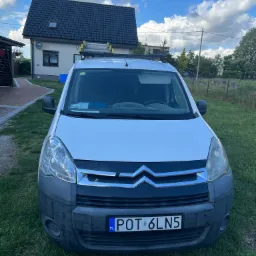 Biały samochód dostawczy Citroen Berlingo z drabina na dachu zaparkowany na trawniku przed domem z ciemną dachówką, sugerujący usługi elektryczne.