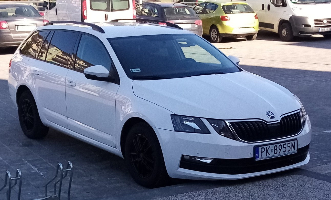 Biała Skoda Octavia kombi z czarnymi felgami zaparkowana na parkingu, widoczna z przodu i boku, z innymi samochodami w tle.