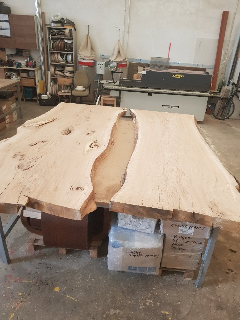 Dwa duże, jasne blaty drewniane z naturalnymi krawędziami, ułożone obok siebie na metalowym stole w warsztacie, z widocznymi kartonami pod spodem i maszynami do obróbki drewna w tle.