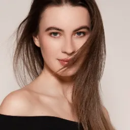 Portret młodej kobiety z długimi brązowymi włosami częściowo zasłaniającymi twarz, ubrana w czarną bluzkę z odkrytymi ramionami na jasnym tle.