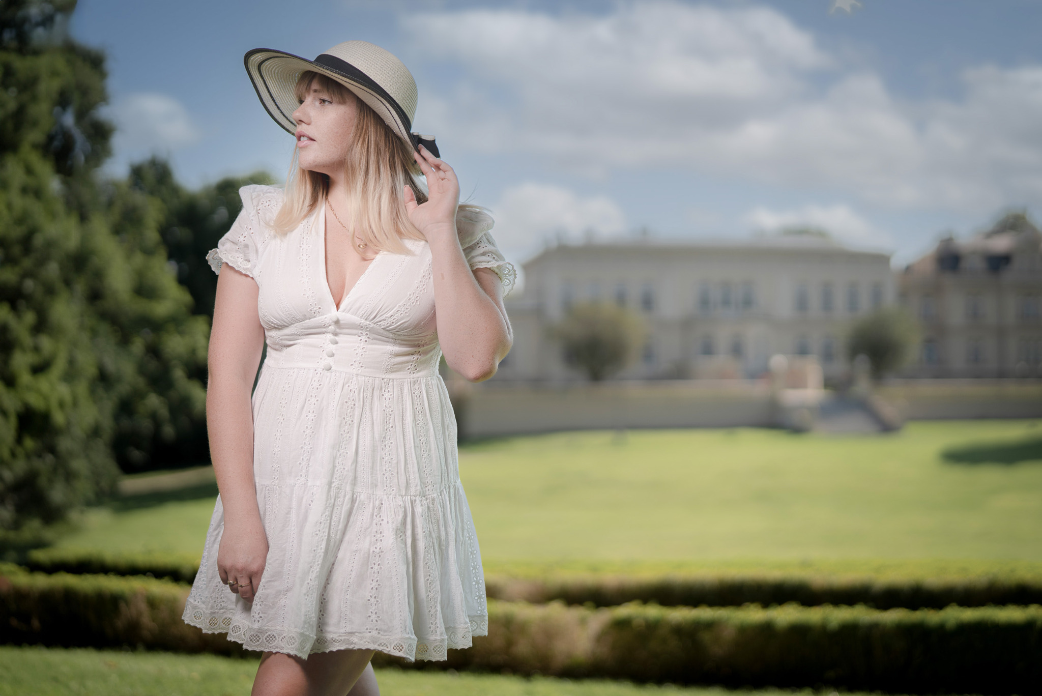 Kobieta w białej sukience i kapeluszu pozuje na tle rozległego trawnika i budynku w oddali, trzymając ręką rondo kapelusza.