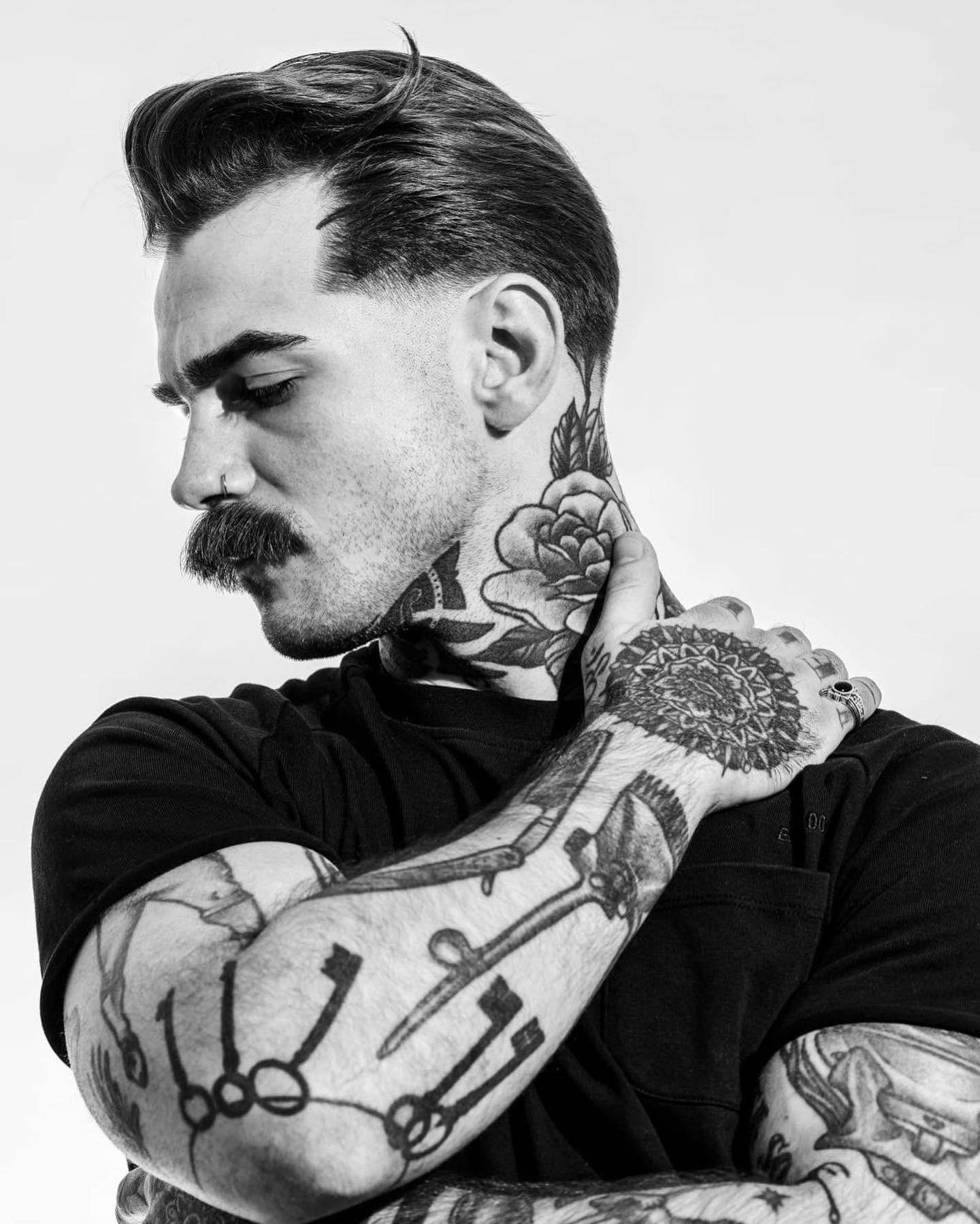 Czarno-biały portret mężczyzny z tatuażami na szyi i ramionach, z wyraźnym wąsem i modną fryzurą, spoglądającego w dół.