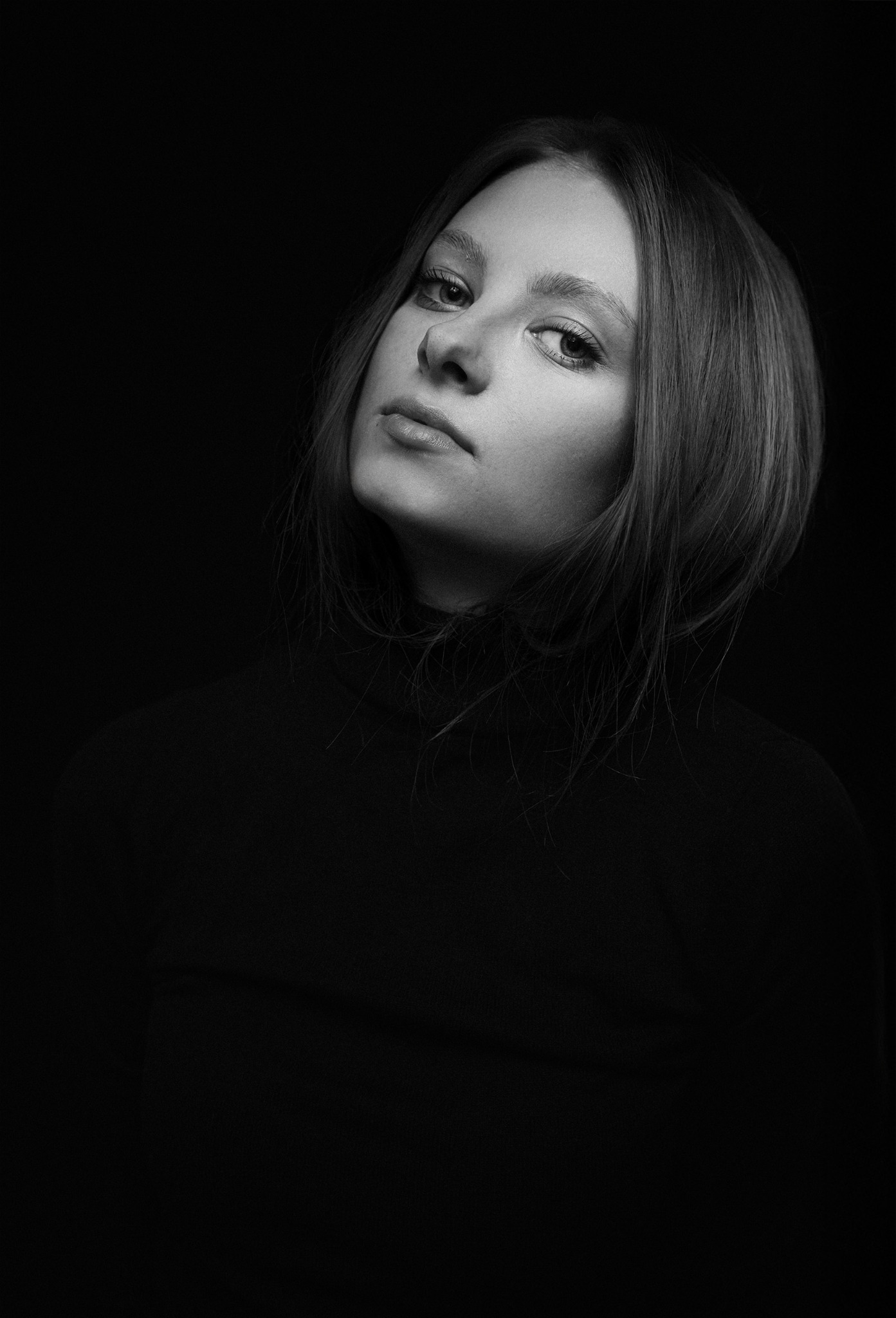 Portret kobiety w czarnym golfie na ciemnym tle, pochylona głowa, artystyczne oświetlenie.