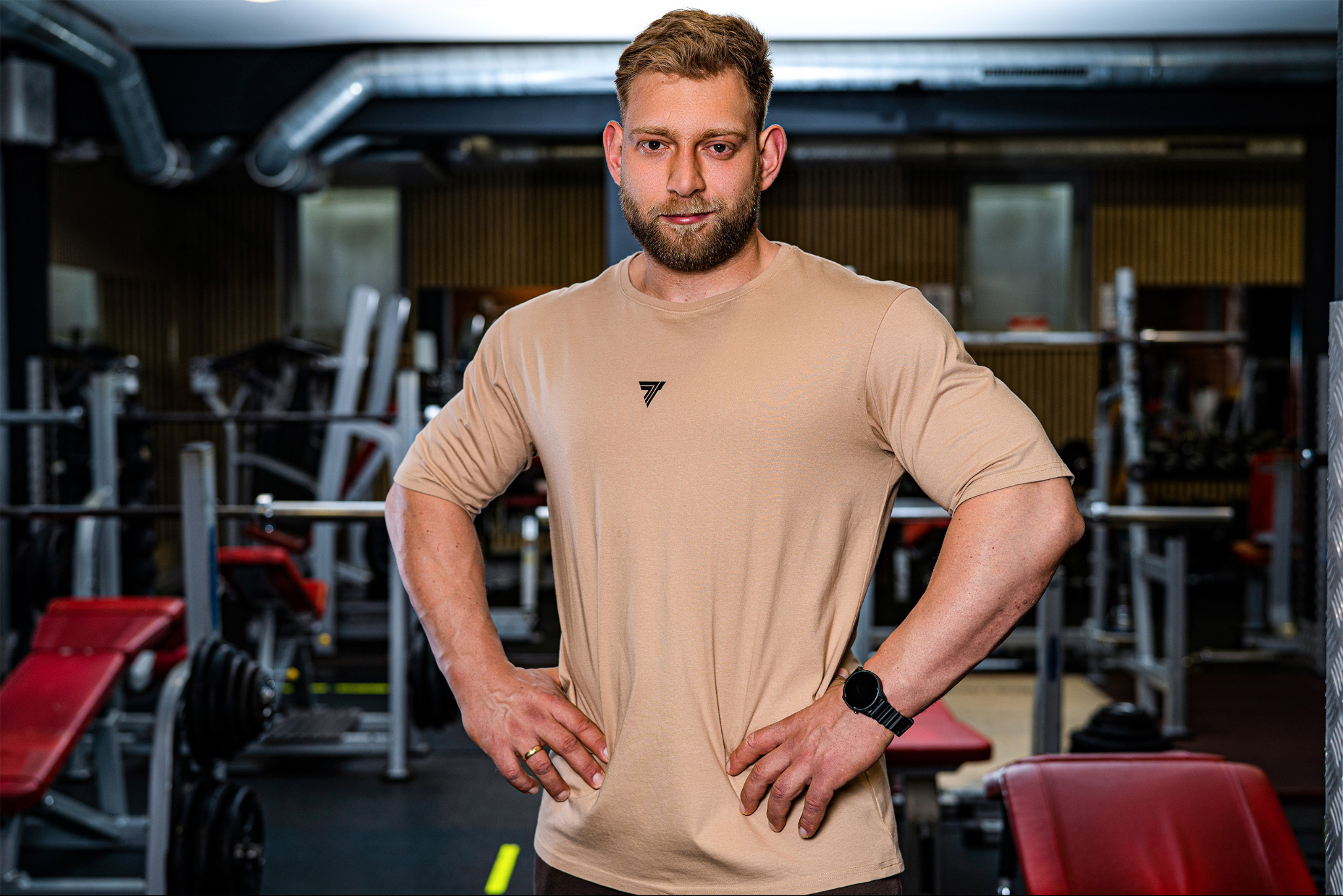 Portret mężczyzny w beżowej koszulce z logo, pozującego w siłowni w Bydgoszczy, z widocznym sprzętem do ćwiczeń w tle.