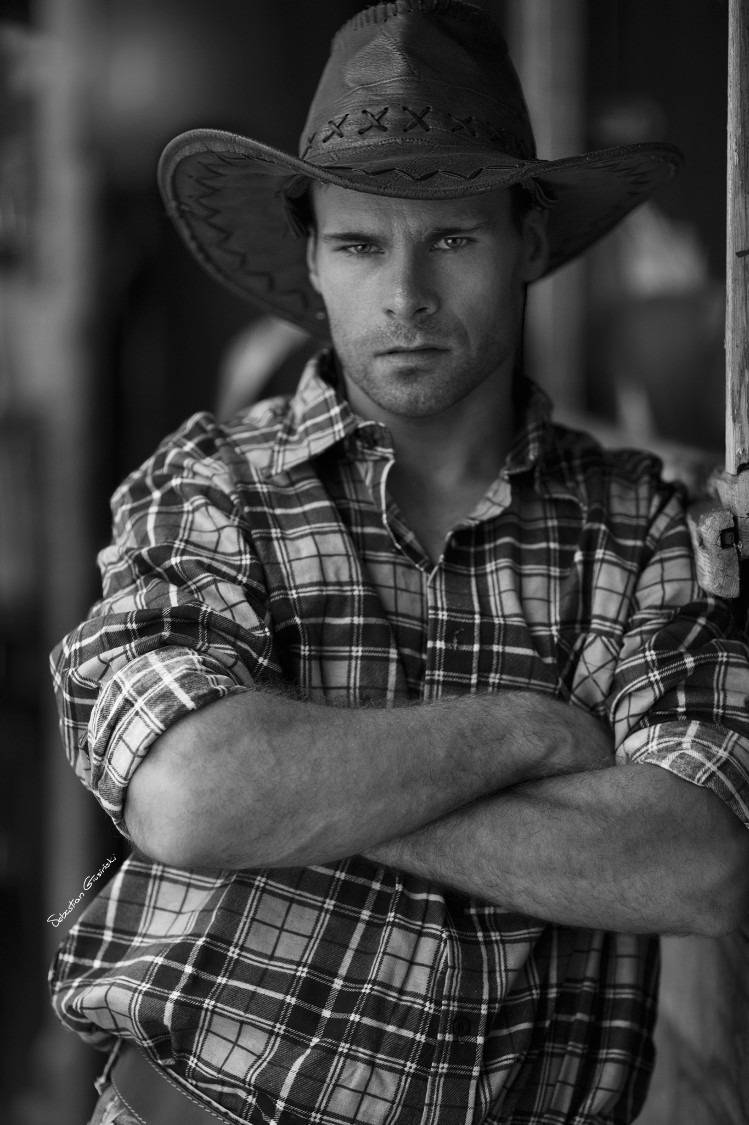 Czarno-biały portret mężczyzny w kapeluszu kowbojskim i koszuli w kratę, z założonymi rękami, oparty o drewnianą ścianę.