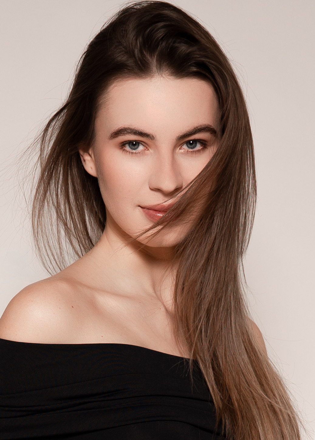 Portret młodej kobiety z długimi brązowymi włosami częściowo zasłaniającymi twarz, ubrana w czarną bluzkę z odkrytymi ramionami na jasnym tle.