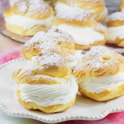 Elegancko ułożone ptysie z kremem, obficie posypane cukrem pudrem na białym talerzu z dekoracyjnym brzegiem, z rozmytym tłem w odcieniach r&oacute;żu i fioletu.