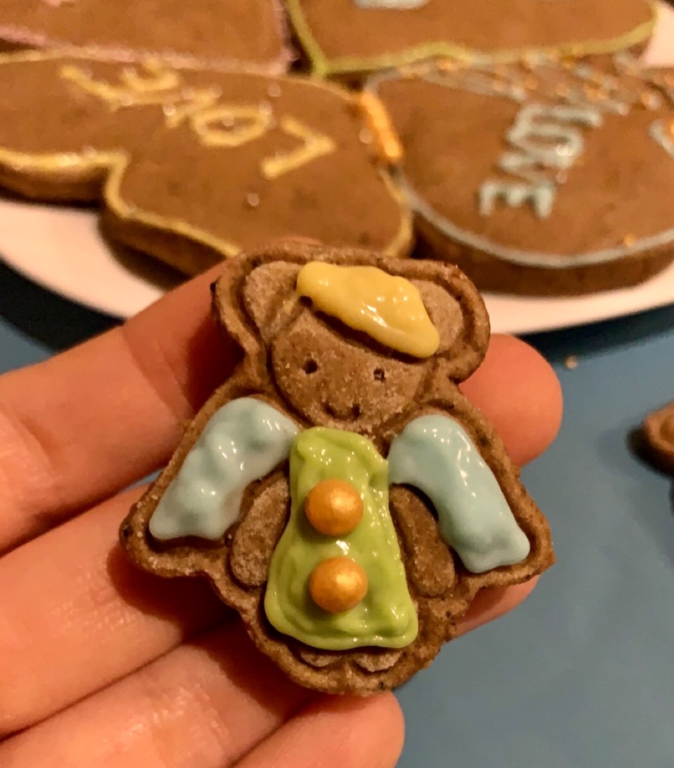 Ręcznie zdobiony piernik w kształcie misia-aniołka z lukrowanymi skrzydłami, aureolą i zieloną tuniką z dwoma złotymi guzikami, trzymany w dłoni na tle innych pierników z napisem LOVE.