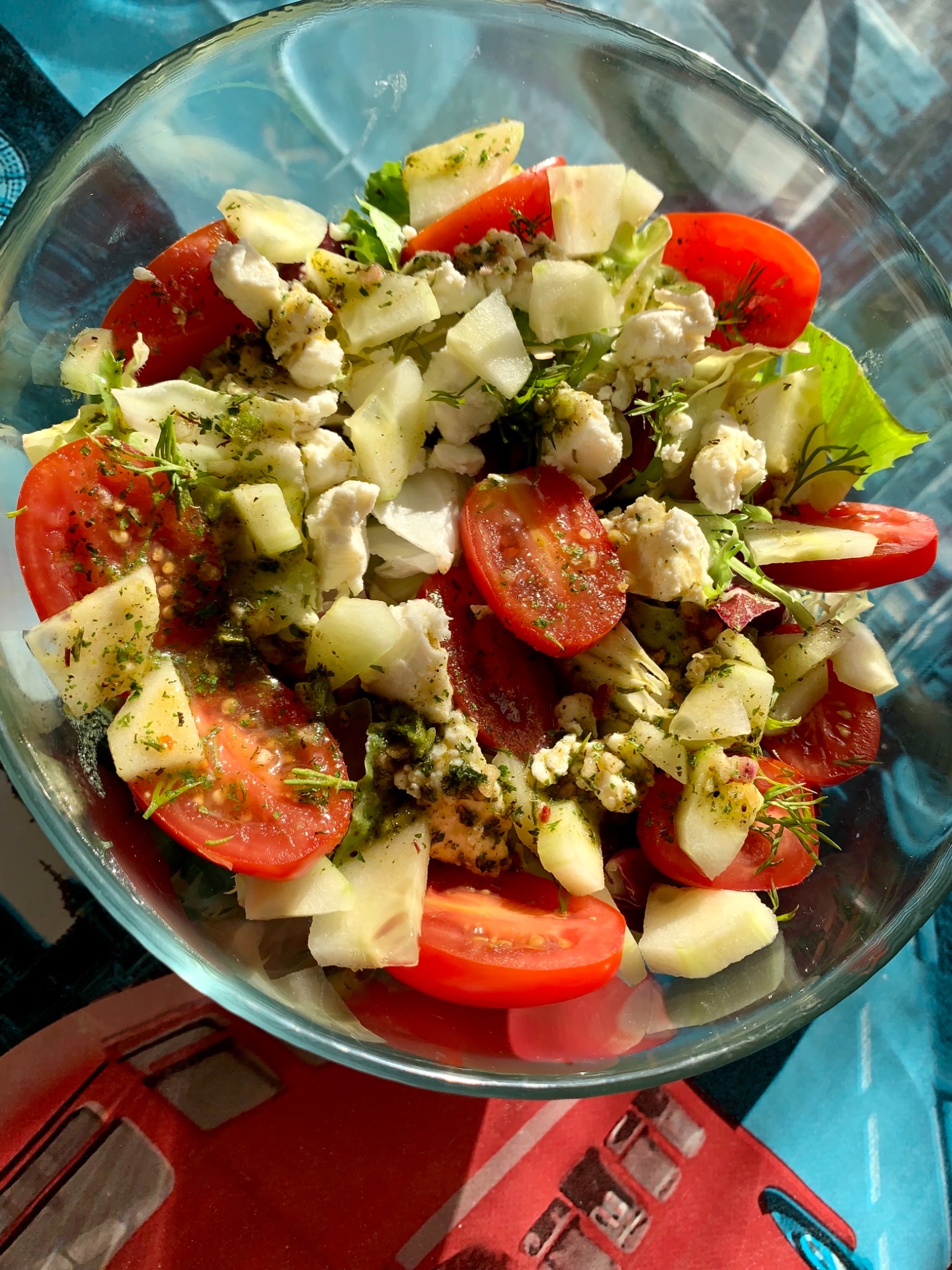 Sałatka ze świeżych pomidorów, ogórków, sera feta i ziół w szklanej misce, widok z góry, na tle wzorzystej serwety.