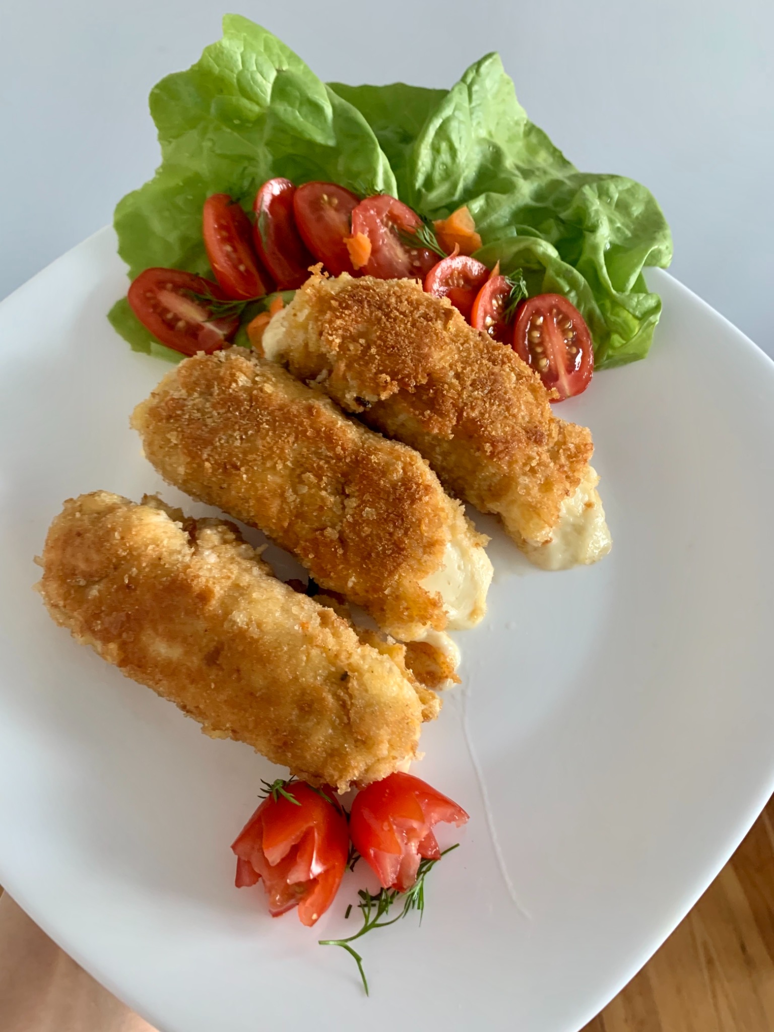 Trzy krokiety z serem w panierce na białym kwadratowym talerzu, udekorowane sałatą, pomidorami i koperkiem.