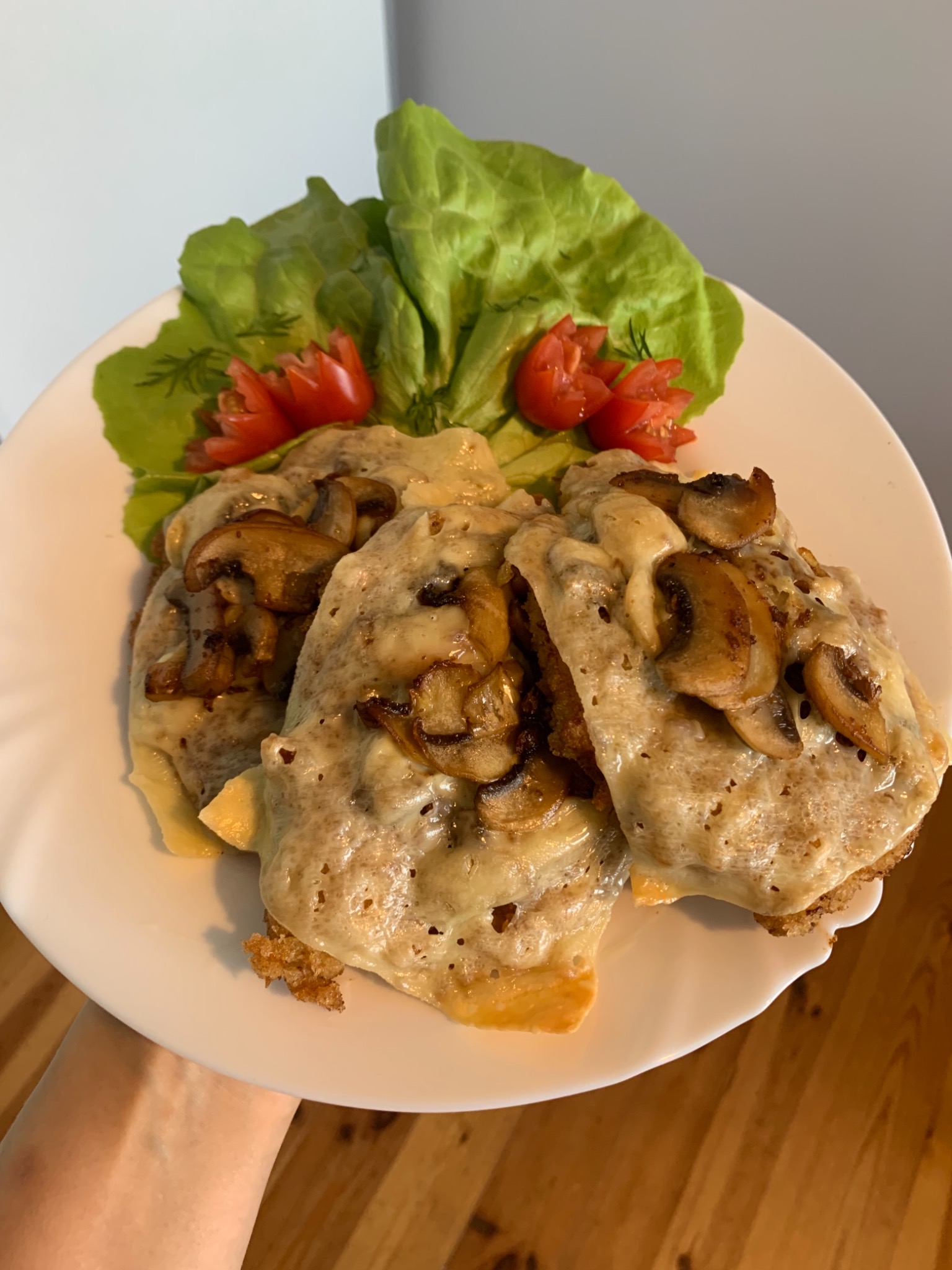 Trzy kotlety w panierce, polane serem i pieczarkami, udekorowane sałatą, pomidorami i koperkiem na białym talerzu, trzymane w dłoni.