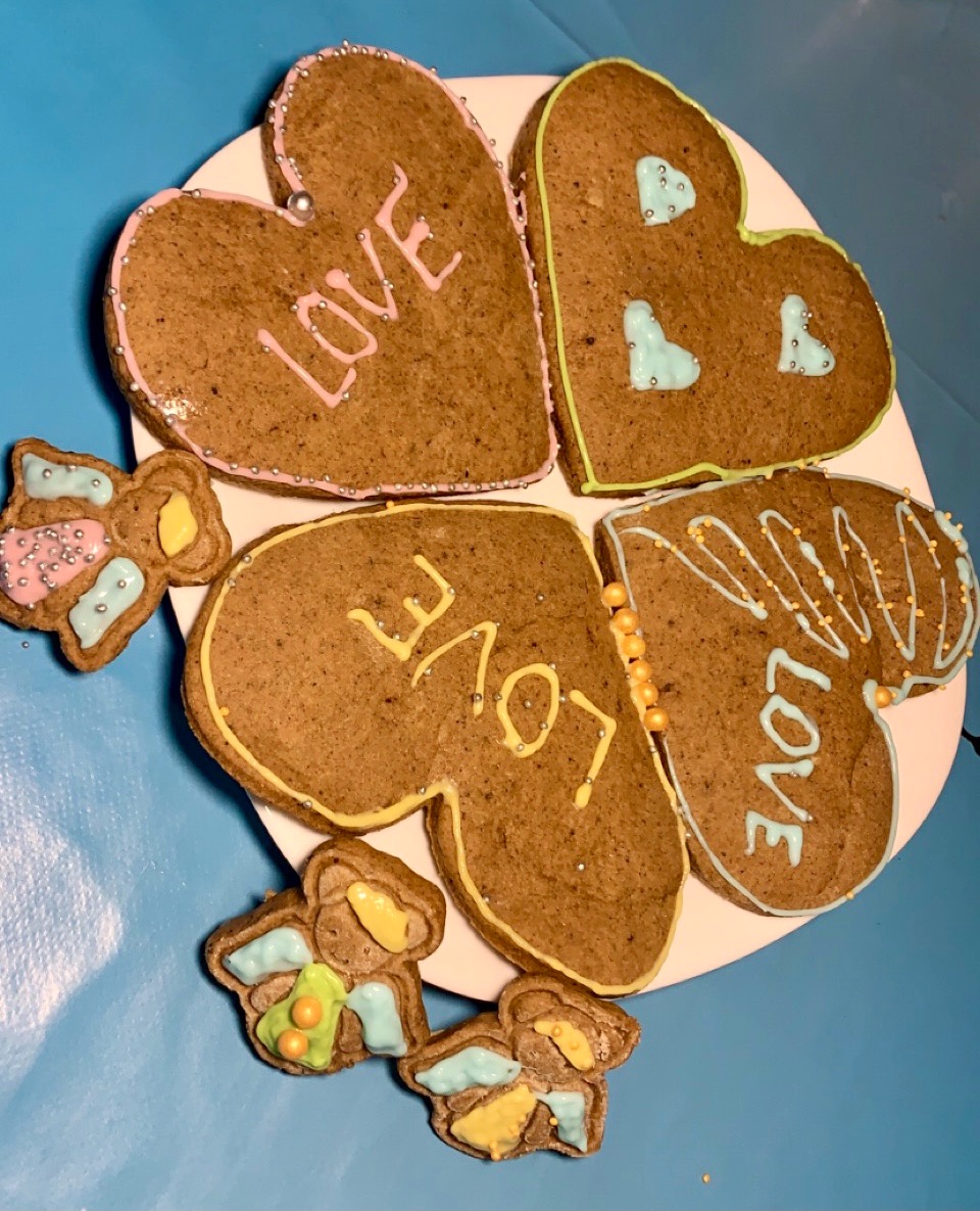 Cztery piernikowe serca ułożone w kształt motyla, udekorowane kolorowym lukrem i napisem 'LOVE', otoczone małymi pierniczkami na białym talerzu, na niebieskim tle.