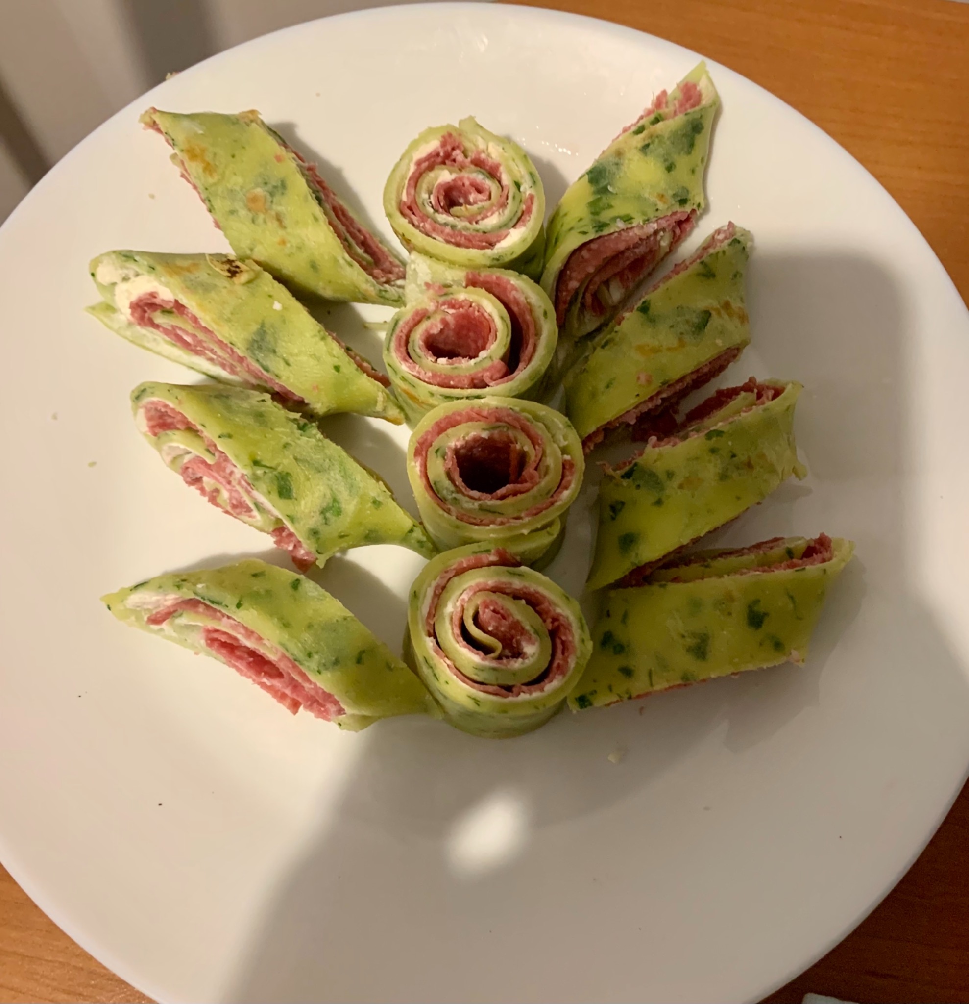 Dekoracyjne roladki z zielonego naleśnika z szynką i kremowym serkiem na białym talerzu, ułożone w centralny wzór rozety.