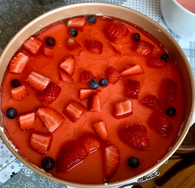 Domowy tort truskawkowy z galaretką i borówkami amerykańskimi w okrągłej formie, widoczny z góry na tle wzorzystej serwetki.