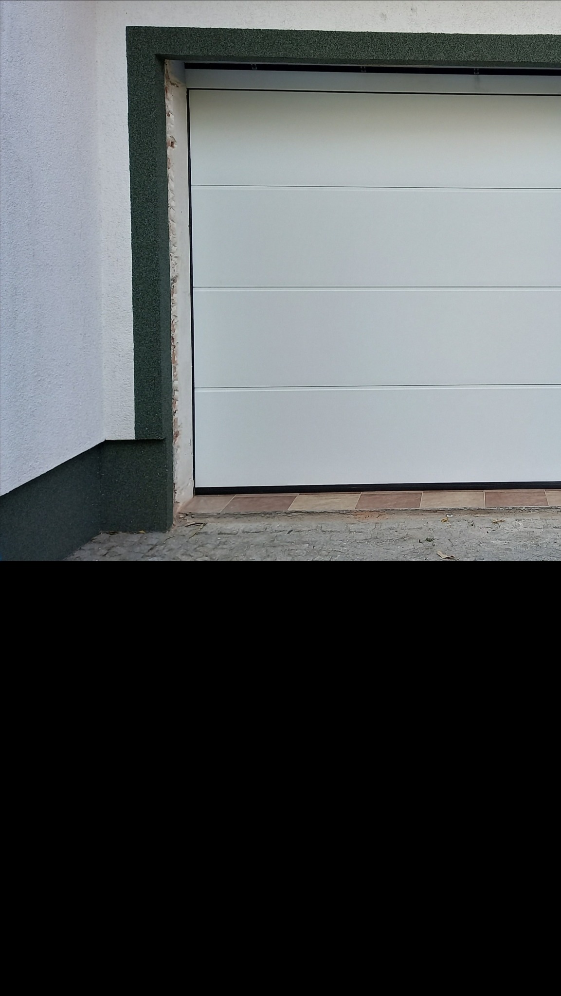 Biała, segmentowa brama garażowa z poziomymi panelami, otoczona szarozieloną ramą, widoczna z zewnątrz na tle białego budynku z szarozielonym cokołem.