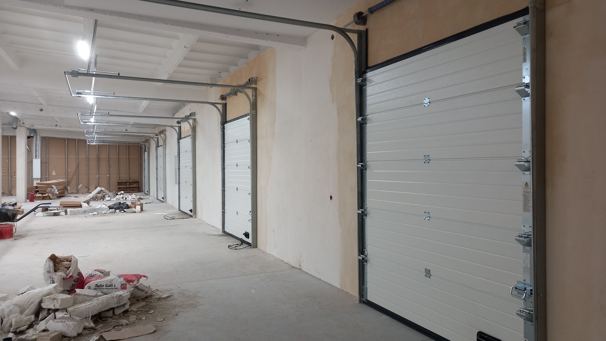 Wnętrze hali z zamontowanymi segmentowymi bramami garażowymi, widoczne ślady prac budowlanych i materiały budowlane.