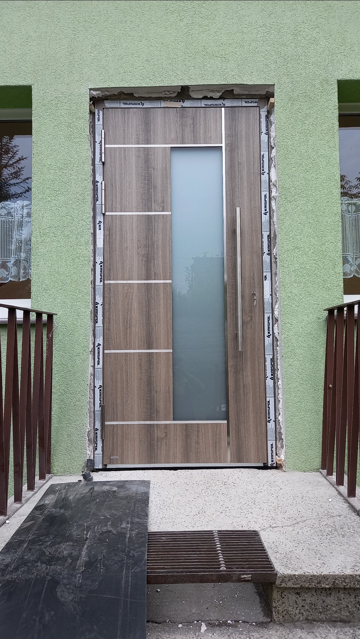 Nowoczesne drzwi wejściowe z drewnopodobnym wykończeniem i szklanym panelem bocznym, osadzone w surowym otworze, widoczne ślady pianki montażowej, metalowa listwa ochronna na futrynie, zielona...