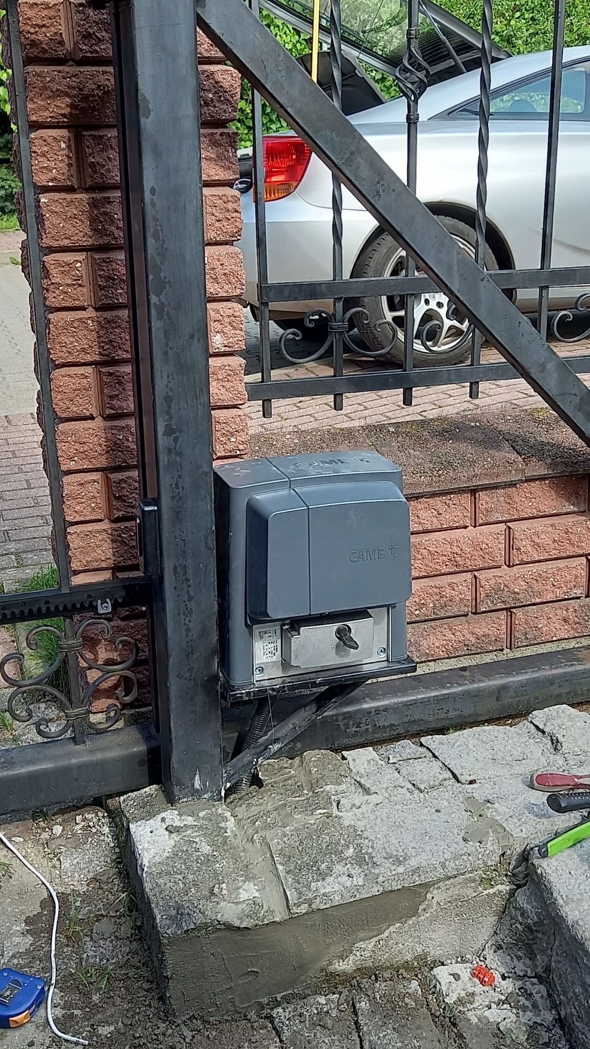 Szary automat do bramy marki CAME zamontowany na czarnej, metalowej bramie wjazdowej, z widocznymi narzędziami i ceglanym słupkiem w tle oraz fragmentem srebrnego samochodu.