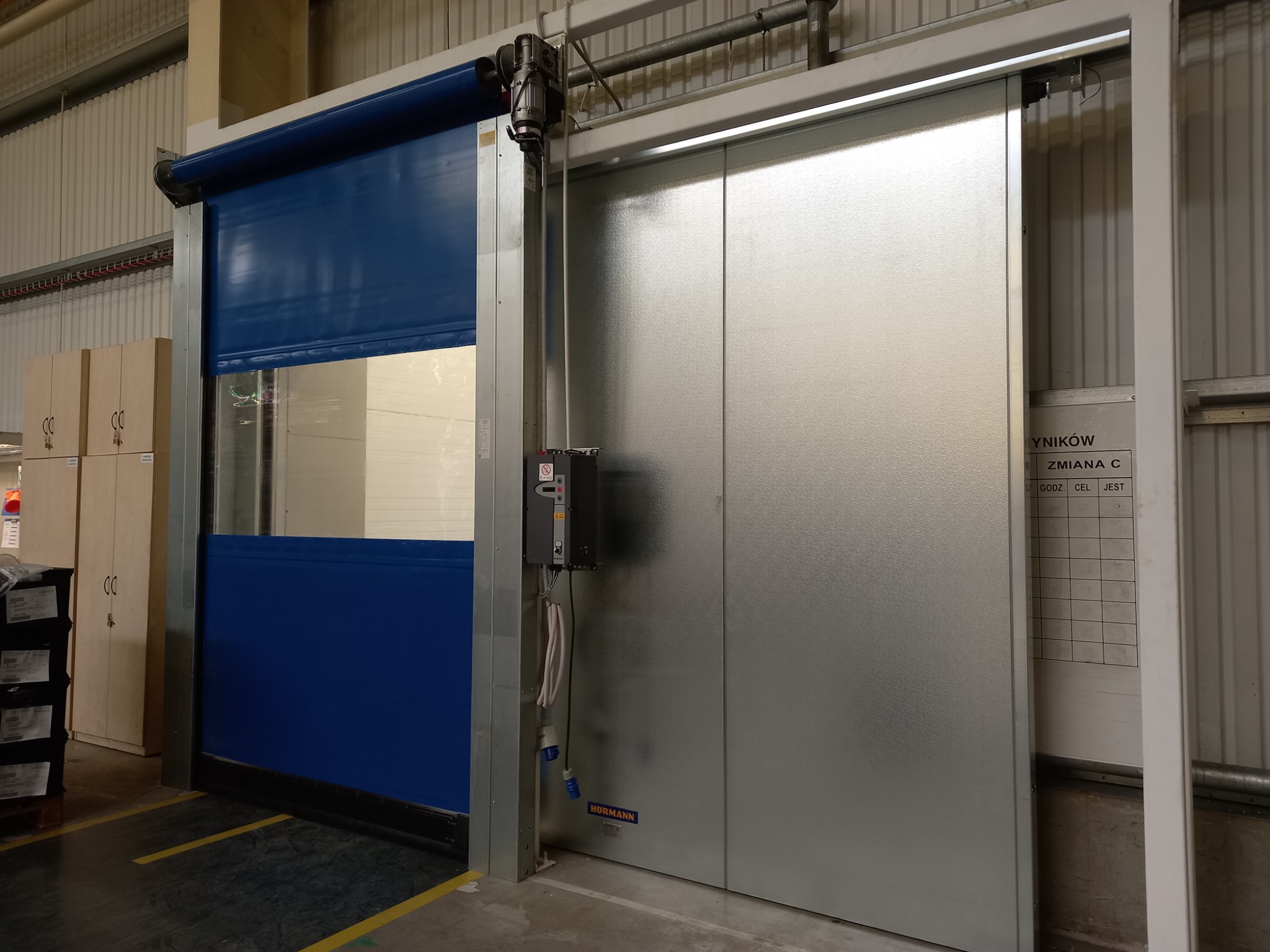 Automatyczna brama przemysłowa: niebieska, rolowana, z okienkiem, po lewej; srebrna, przesuwna, po prawej, z widocznym mechanizmem sterującym i tablicą wyników na ścianie w tle.
