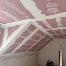 SOBEX TEAM - PATRYK SOBIŚ - Poddasze z płytami kartonowo-gipsowymi, wstępnie wyszpachlowane na łączeniach. Widoczne narzędzia i poziomice. Skos dachu z widocznymi elementami konstrukcyjnymi.