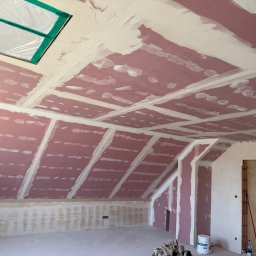 SOBEX TEAM - PATRYK SOBIŚ - Poddasze z płytami gipsowo-kartonowymi w trakcie szpachlowania, widoczne łączenia i fragment okna dachowego zabezpieczonego folią. Ściany częściowo wyszpachlowane.