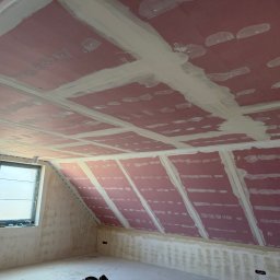 SOBEX TEAM - PATRYK SOBIŚ - Poddasze z oknem po montażu płyt gipsowych i wstępnym szpachlowaniu łączeń. Widoczne różowe płyty i białe pasy szpachli. Ściany częściowo przygotowane do dalszych prac.