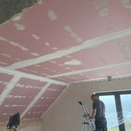 SOBEX TEAM - PATRYK SOBIŚ - Montaż płyt gipsowo-kartonowych na poddaszu. Widoczny pracownik na drabinie, różowe płyty z nałożoną białą masą szpachlową. Okno zabezpieczone folią.