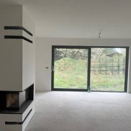 Sobex Team - Wnętrze po remoncie: białe ściany, surowa podłoga, nowoczesny kominek z czarnymi elementami i duże okno z widokiem na zieleń. Minimalistyczny design.
