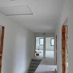 Sobex Team - Wnętrze w trakcie wykańczania: białe ściany, sufit z otworem rewizyjnym, widoczne instalacje elektryczne w murze, schody i narzędzia w tle. Jasne, naturalne światło.